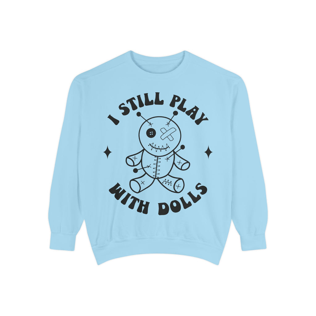 A light blue crewneck sweatshirt featuring a black graphic of a stitched voodoo doll with the text 'I STILL PLAY WITH DOLLS' arched above and below it, displayed on a clean white background.