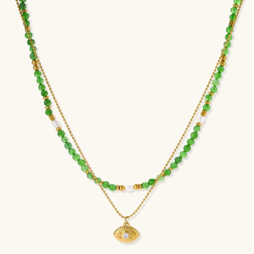 Layered necklace with green stone beads, gold spacers, white accents, and protective eye pendant on cream background.