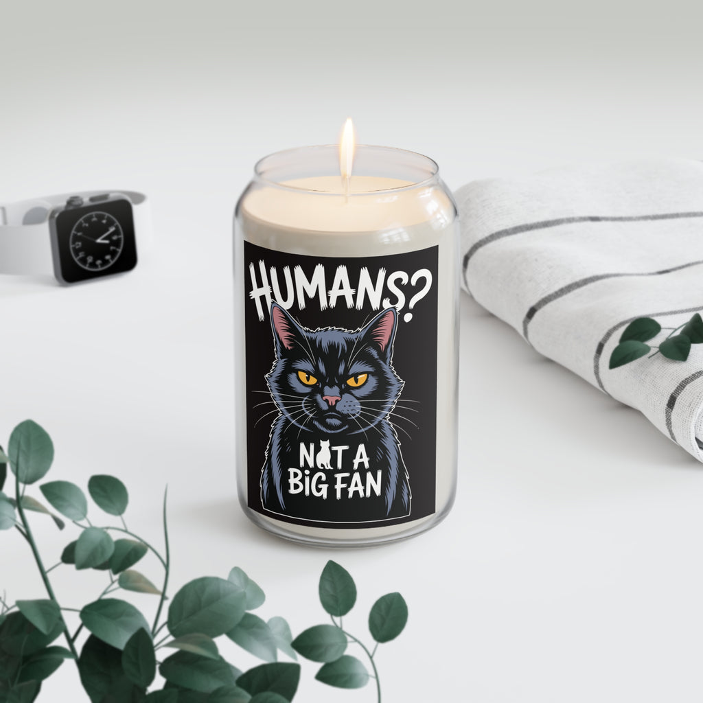 Candle with a black cat illustration and text 'Humans? Not a big fan' on a white surface with a plant and watch in the background.