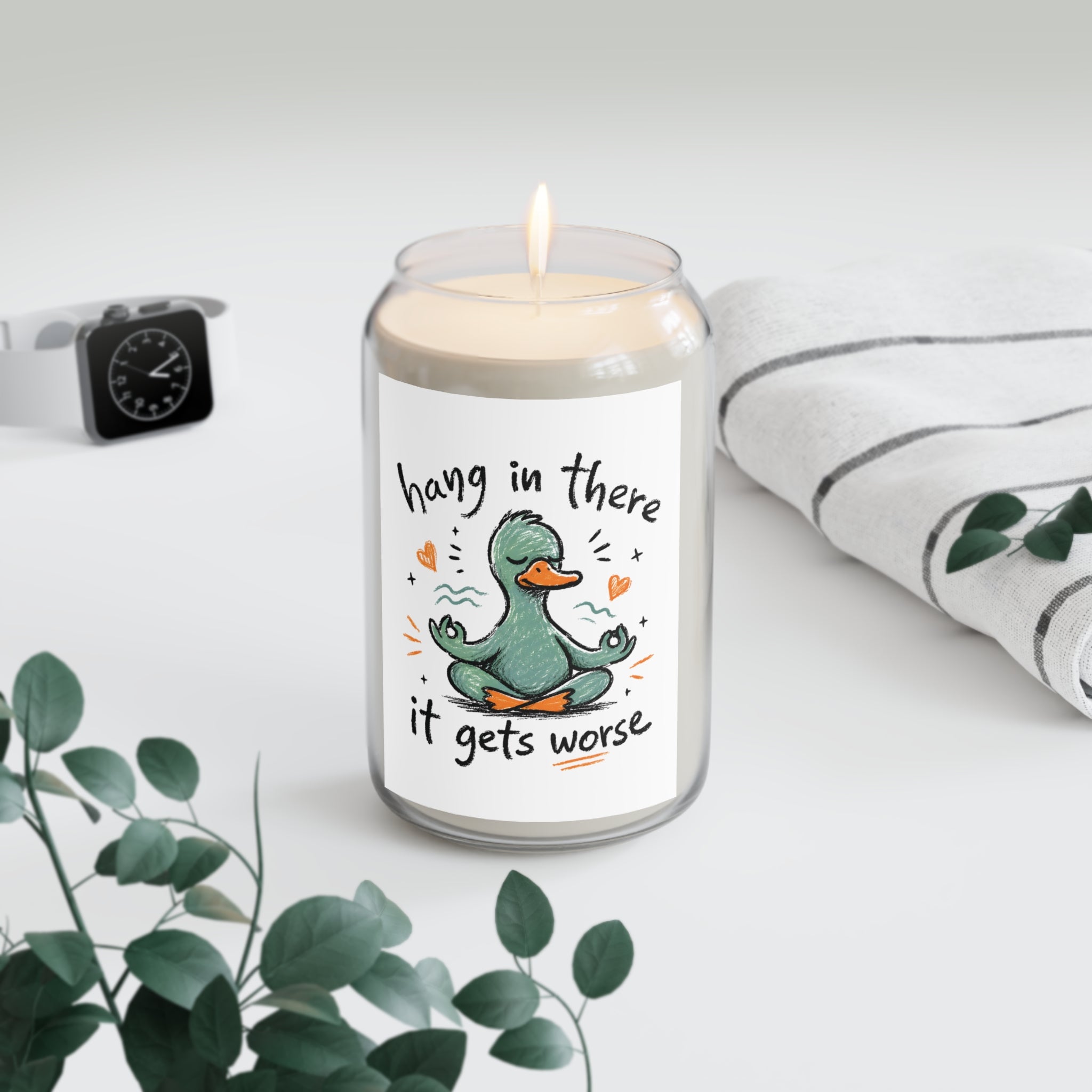 Candle in a glass jar with a duck illustration and text on a white surface with a plant and watch in the background.